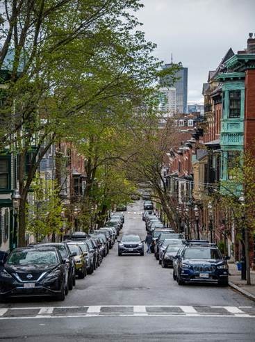 Tree-lined residential street in a Greater Boston neighborhood with parked cars, historic brownstones, and the city skyline in the background.