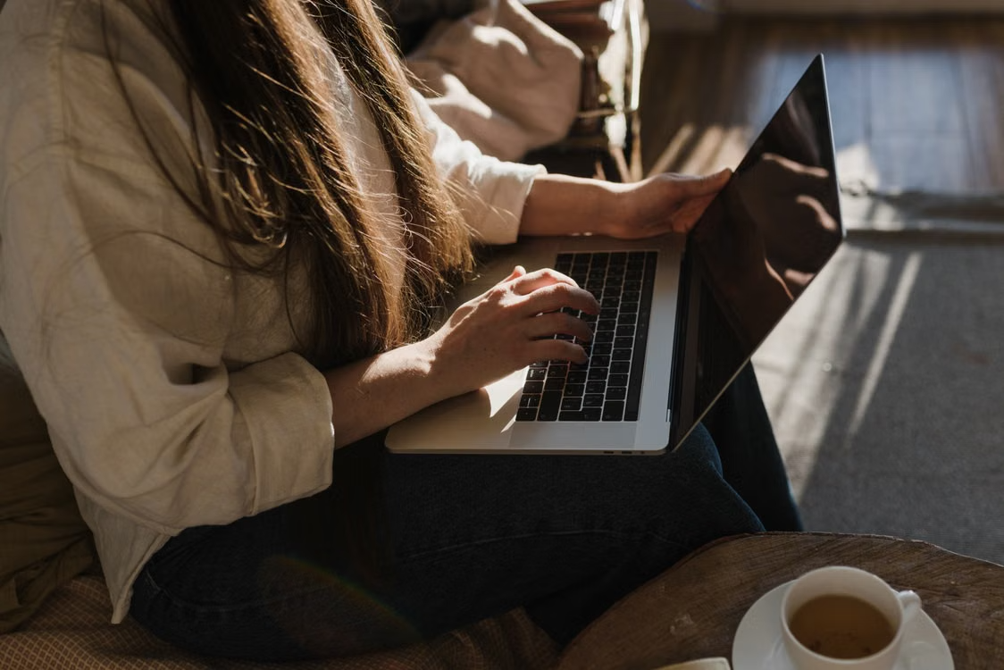Close-up of a woman using a laptop in a warm, cozy indoor setting with sunlight. Courtesy of Pexels